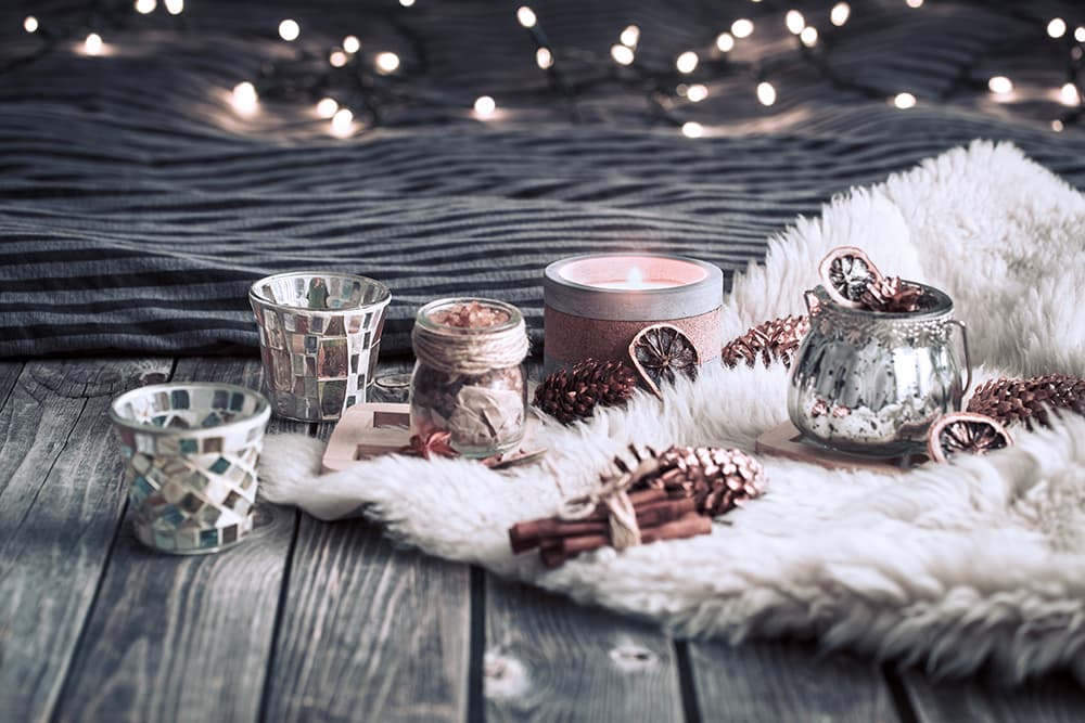 Meleg hangulatú téli dekoráció: égő gyertyák, díszes mécsestartók, fahéj és tobozok egy fehér szőrmén, karácsonyi fényfüzérrel a háttérben