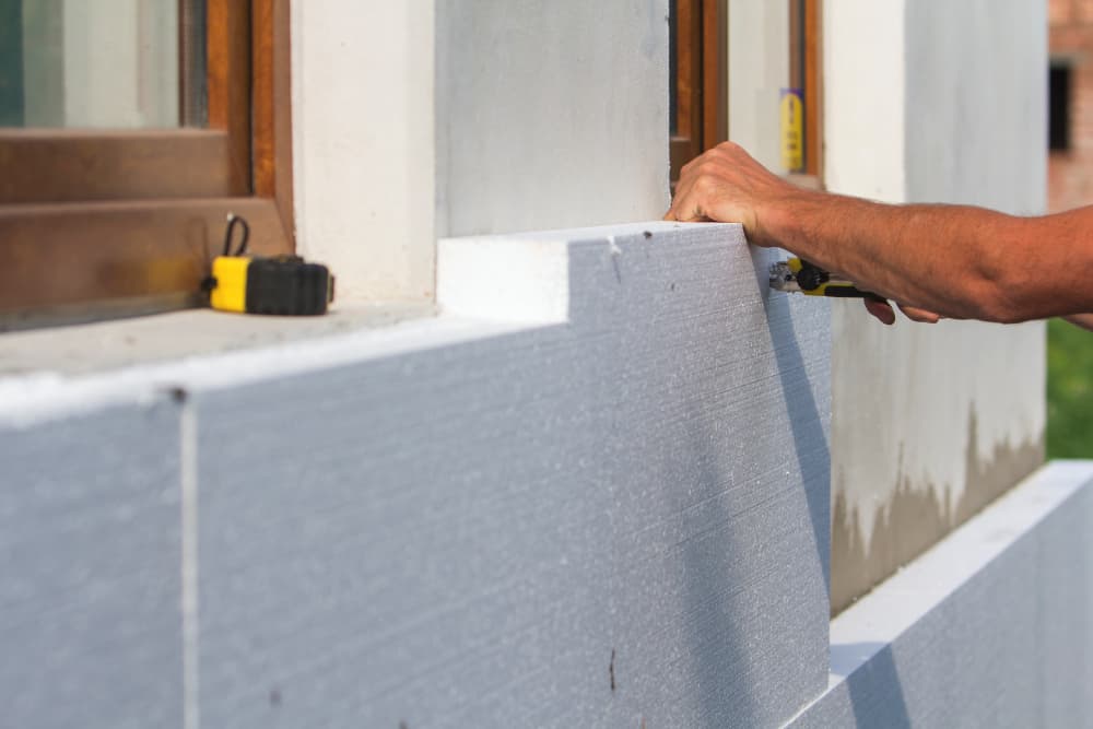 Egy szakember keze, amint egy sniccerrel pontos méretre vágja a fehér hőszigetelő lapot egy ablakkeret melletti részen.