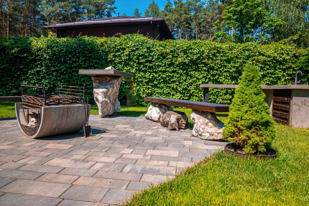 Modern, minimalista pihenőhely a kertben, egyedi beton tűzrakóval, kőből és fából készült padokkal, szürke térkő burkolaton, zöld sövény előtt.