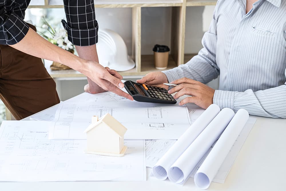 Két szakember tervrajzok fölött számológéppel egyezteti egy felújítás vagy építkezés költségeit