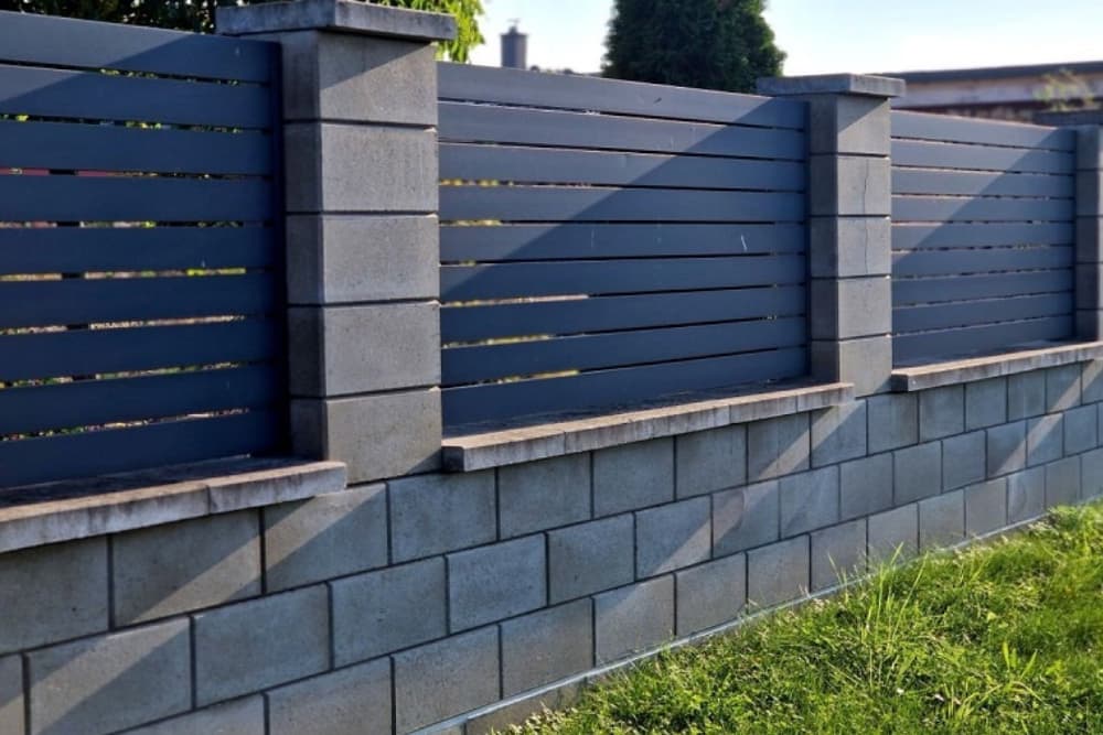Modern kerítés szürke betonelemekből és sötétszürke fém lamellákból, zöld gyep mellett.