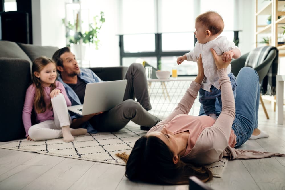 Egy négytagú család pihen a padlón egy nappaliban, ahol az anya felemeli a babáját a levegőbe, míg az apa és a lánya hátulról nézik őket.